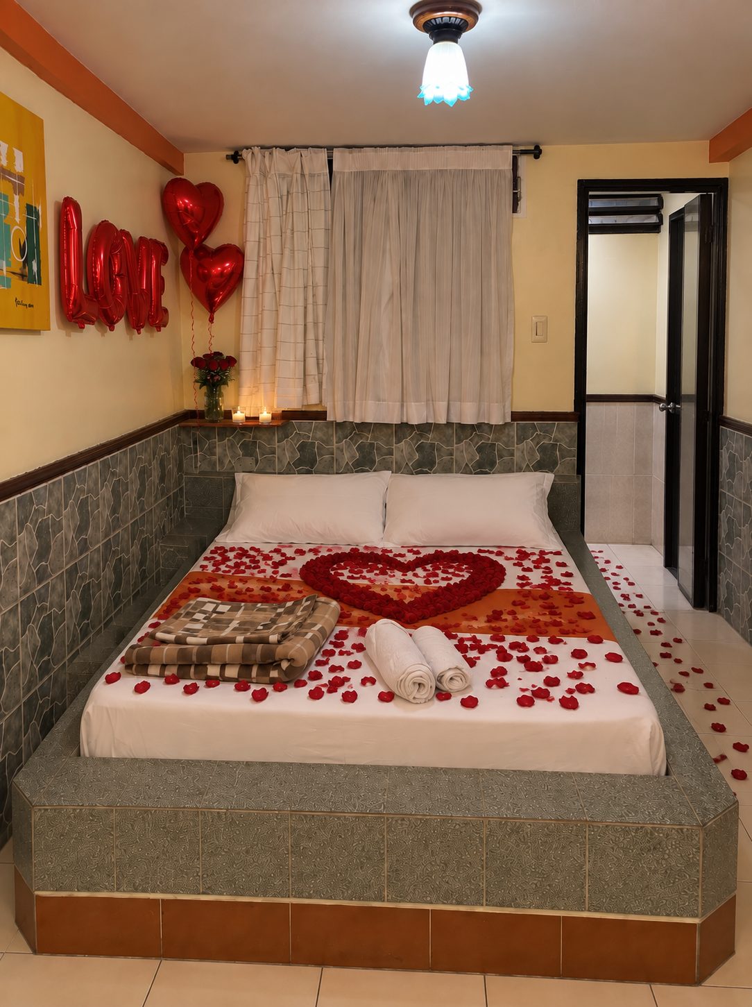 Cama matrimonial decorada con corazón de pétalos de rosa, globos LOVE y velas